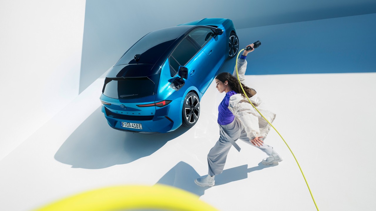 Vue de dessus d'une Opel Astra Electric bleue à toit noir avec une femme faisant tourner un câble de recharge jaune.