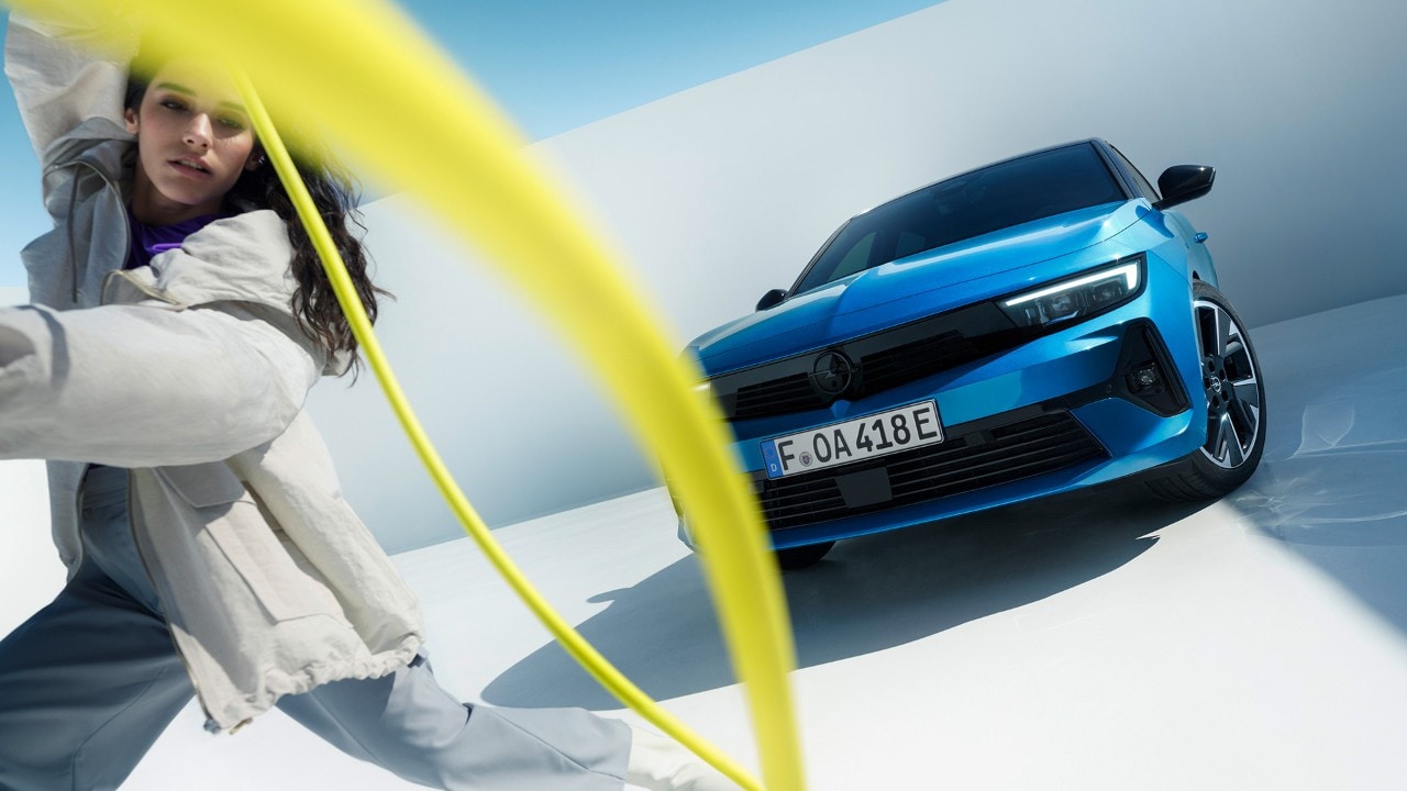 Vue de face d'une Opel Astra Electric bleue avec une femme faisant tourner un câble de recharge jaune.