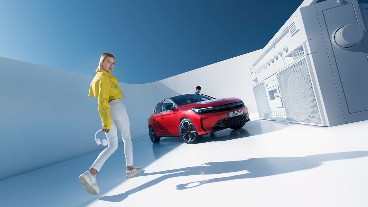 Femme debout à côté de la nouvelle Opel Corsa de couleur rouge avec toit noir et un homme montant par la porte conducteur.